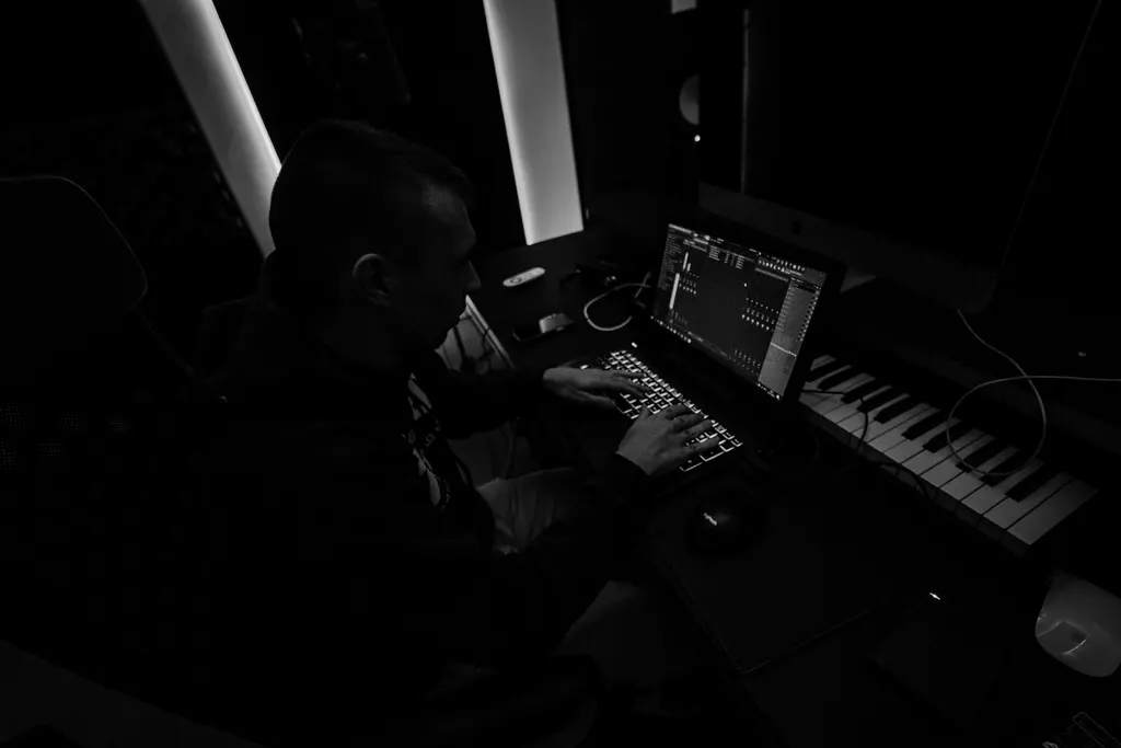Un produttore musicale compone al computer in uno studio in penombra, con una tastiera davanti.